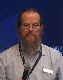 A color photograph of a man in glasses with thinning reddish-brown hair and a full beard, holding a microphone and wearing a name badge.