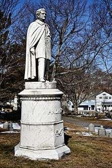 A white marble statue of a man wearing 19th century clothing and a long cloak stands atop a tall marble pedestal. The statue stands on a small grassy knoll with bare tree branches and a blue sky in the background.