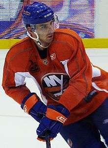 Hockey player in orange uniform. He skates across the ice, holding his stick, and looks over his left shoulder.