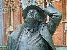 A bronze statue of an old man, wearing a suit and tie and an open long coat, looking up and holding a hat on his head with his left hand; behind, part of a brick building with Gothic arched windows