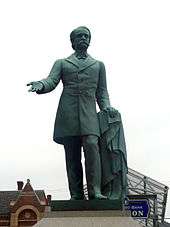 A statue depicting a mustachioed man holding an overcoat in his left hand with his right hand extended