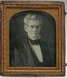Serious look, black suit and bow tie with white shirt, full profile. Image is in a gold frame.