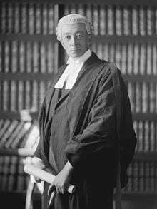 man in his early 50s wearing a lawyers' wig and gown, standing in front of a book shelf