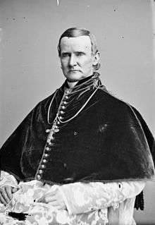 A black and white photograph of a man with short dark hair wearing a dark buttoned garment around his beck and upper chess with a cross. Below it his lap and robes are visible.