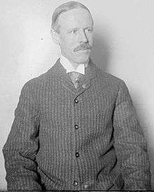 White man in medium shot.  Man has receding hairline and mustache.  He is wearing a collared shirt and a button down sweater or pinstriped jacket.