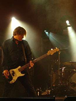 Collins is playing his bass guitar with his left hand on the fret board and right hand with fingers plucking at the strings. His head is bent to his left and down, his fair haired fringe hangs over his eyes. He wears a dark long shirt. A drum kit is beyond him, further to his left. Stage lights shine down from above.