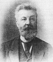 Head and shoulders of a bearded man wearing a three-piece suit, tie and high collar