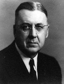 An aging, unsmiling man with round-rimmed glasses