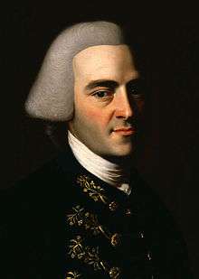 Half-length portrait of a man with a hint of a smile. His handsome features suggests that he is in his 30s, although he wears an off-white wig in the style of an English gentleman that makes him appear older. His dark suit has fancy embroidery.