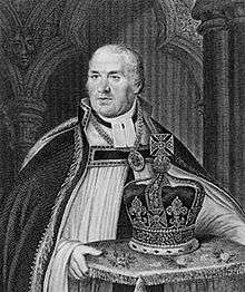 A middle-aged man wearing clerical robes and gown carrying a decorated cushion, upon which rests a large bejewelled crown