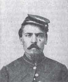 Head of a white man with a Van Dyke mustache and goatee, wearing a cap tilted on his head and a military jacket buttoned only at the neck.