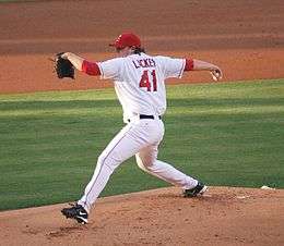 A man in a white baseball uniform with red trim throws a baseball from a dirt mound on a grass field. Under his jersey, he is wearing a long-sleeved red shirt with the sleeves pushed up to his elbows. He wears a black baseball glove on his right hand and a red baseball cap on his head.