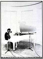 A black and white photo of Lennon sitting at a white parlour grand piano. He is wearing headphones and a dark shirt.