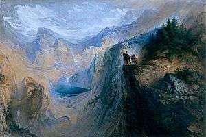 Two men stand on a cliff overlooking a valley surrounded by forests and tall mountains.