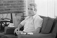 White-haired man in thirties sitting in a chair, pack of cigarettes readily available
