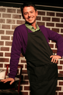 A grinning man in front of a faux-brick wall wears a purple shirt under a black apron while leaning with his right arm on a low table