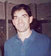 Head shot of a man, wearing a blue shirt, stands in a locker room