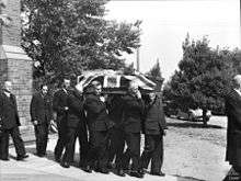 Black and white photo of a coffin which has been draped with a British flag being carried by pallbearers. The pallbearers are middle-aged men wearing formal suits, and three other middle aged men in suits are following the pallbearers.