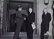 Black and white photograph of a man wearing military uniform shaking the hand of a man in a formal suit. The two men are standing in front of a stone gateway, and another man wearing a formal suit is looking on from the right of the image.