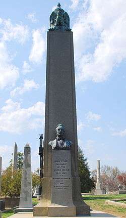 A large obelisk in a graveyard, with a bust of Tyler, and a black cast iron cage partially visible behind it.