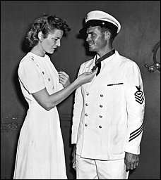 A woman stands next to a man dressed in chief petty officer dress whites admiring a medal of honor the man is wearing