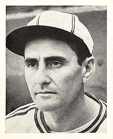 A man in a white baseball jersey and cap with dark stripes looks to the left.
