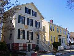 Two houses stand near the street, with no front yard. The left house is a two story white colonial, and the right one is also a two story colonial, painted yellow.