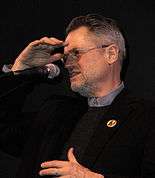 A man wearing a black coat and sweater over a grey shirt is seen with his right hand over his face and microphone in front of him.