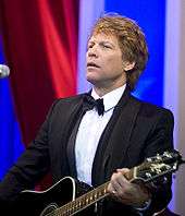 Jon Bon Jovi, a caucasian male in his mid-40s with dark blond hair, wears a black suit and white shirt with a black bow-tie. He is seen with a black acoustic guitar strapped across his stomach.