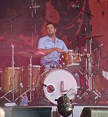 Coghill is playing his drum kit, his right arm is crooked, holding a drumstick and hitting the drum rim situated slightly to his left. His left arm and most of his lower body is obscured by the rest of the kit. He stares off to his right with his lightly bearded chin slightly raised. He is dressed in a pale blue sleeveless shirt and similar coloured pants. In front and above his kit are microphones and band equipment. Behind him is a large graphic design, consisting of red-orange lights and black dots.