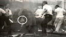low-resolution monochrome image showing several people in a pub, scattering; one is a policeman. One man, his face circled, sits on the floor in front of the bar, looking bemused.
