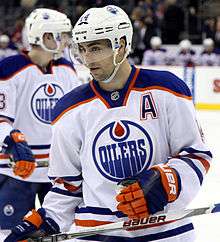 A Caucasian ice hockey player wearing a white jersey with a blue and orange circular logo with the word "OILERS".
