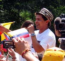 A man with a hat and a white t-shirt singing to a microphone.