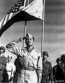 Filipino male in World War II Army Khaki uniform saluting in the foreground. A color guard in the background; the 48-star United States flag on the viewers left, a reversed/war Philippine Commonwealth flag on the viewers right.