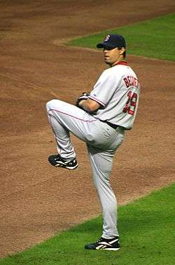 A man in a gray baseball uniform with red trim stands on a dirt-and-grass field with his left leg lifted. He is wearing a black baseball glove on his left hand, and looking over his left shoulder.