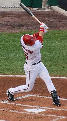 A man, clutching a baseball bat and wearing a red batting helmet and a white baseball uniform with his surname and number partially obscured on his back, swings at a pitch.