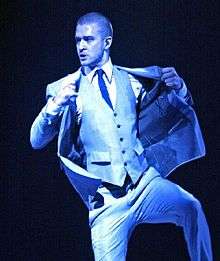 A man with blue light being cast down upon him. He is wearing a suit, including a vest and tie, and his hands are placed on his jacket.