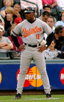 A dark-skinned man in his mid-forties wearing a gray baseball uniform stands with arms akimbo. His uniform reads "Orioles" in orange script lettering across the chest, with a block "11" below it in orange, and is accompanied by a black batting helmet.