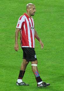 A photograph of a bald man wearing a red and white striped shirt, black shorts and black socks.