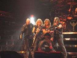 Four men standing next to one another on a stage, three of which are holding guitars. All four men are wearing black clothing, and some of the articles of clothing are studded.
