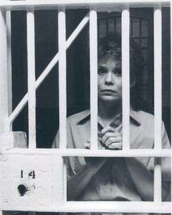 A woman with short hair, wearing shirt, while holding the bars of a fictional prison.