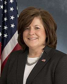 A female Caucasian wearing a blue suit with a white blouse and an American flag lapel pin. In the background is an American flag.