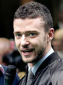 Head and shoulders of Justin Timberlake, wearing a light blue shirt, ablack and white tie and a black jacket, speaking into a microphone