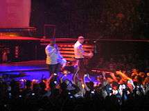 In a stage, two men, both wearing white shirts, can be seen holding microphones. The first man, wearing a long sleeve shirt, sings, while the other man, wearing a shirt, dances to the music played. A crowd of people can also be seen holding up their cellphones.
