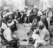 An old photo of a dozen old and middle-aged men sitting on the ground around a mat. A man in front sits next to a mortar and holds a bat, ready for grinding. A man opposite to him holds a long spoon.