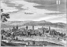 Blank and white print of a 17th-century city showing a city wall, a large cathedral and a large public building. In the background are mountains.