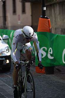A road racing cyclist wearing a mostly white skinsuit with black, green, and gold trim crouches low on his bicycle while riding out of the saddle. A car following behind him and a roadside barricade are also visible.