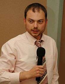 A standing bearded Russian man while wearing a pink shirt and striped necktie while holding a wireless black microphone; on his right index finger is a shiny golden wedding ring.