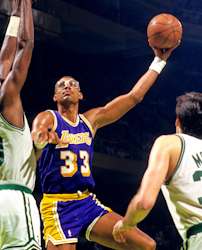 A man, wearing a purple jersey with the word "LAKERS" and the number 33 on the front, is trying to shoot the basketball while being guarded by two man who are wearing white jerseys.