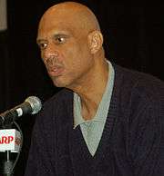 Bald headed black man speaking into a microphone. Headshot only with a polo shirt and sweater.
